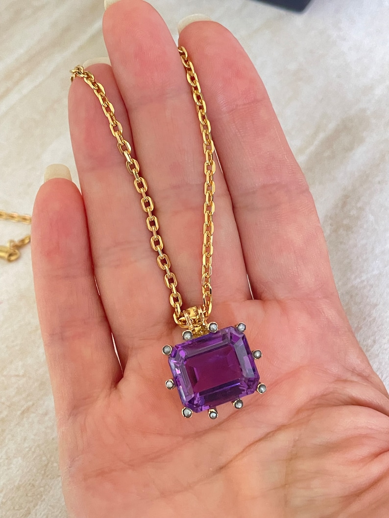 May include: A gold chain necklace with a large, rectangular, purple amethyst pendant. The pendant is set in a gold frame with small, white pearls around the edges.