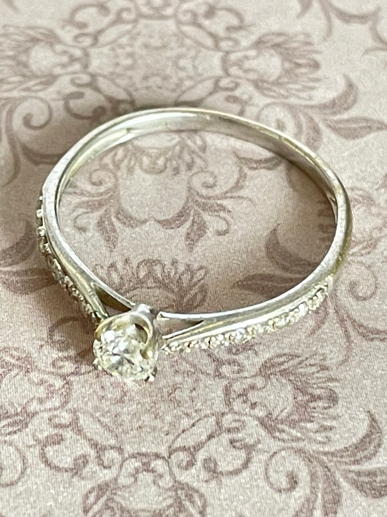 K&ouml;nnte beinhalten: Ein zarter Silberring mit einem einzelnen runden Diamanten in der Mitte und kleineren Diamanten, die entlang des Bandes gesetzt sind.
