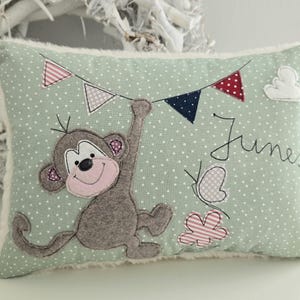 Könnte beinhalten: Ein dekoratives Kissen mit hellgrünem Hintergrund und weißen Punkten. Es zeigt einen applizierten Affen, der an einer Girlande aus bunten Wimpeln hängt. Der Name "Junes" ist zusammen mit einem Schmetterling und einer Blume aufgestickt.