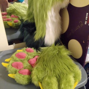 May include: A green and white faux fur headpiece with long ears and matching green paw gloves featuring pink paw pads and yellow claws. The headpiece has a white chest section and dark green accents, while the gloves are covered in long green fur.
