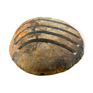 May include: A round, weathered stone with horizontal grooves. The stone has a mix of brown, orange, and black hues, suggesting natural aging and texture. The grooves are dark, contrasting with the lighter stone.