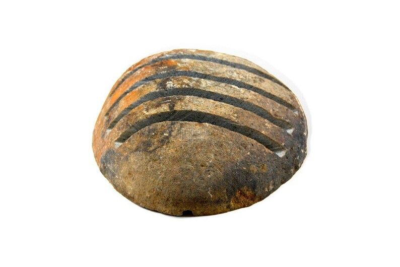 May include: A round, weathered stone with horizontal grooves. The stone has a mix of brown, orange, and black hues, suggesting natural aging and texture. The grooves are dark, contrasting with the lighter stone.