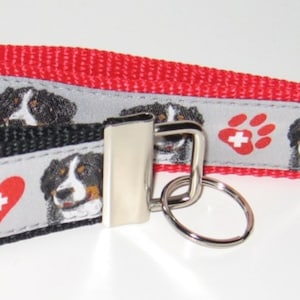Könnte beinhalten: Zwei Schlüsselanhänger mit einem Berner Sennenhund-Design. Ein Schlüsselanhänger ist schwarz mit einem grau-weißen Streifen und einem roten Herzen mit einem weißen Kreuz. Der andere Schlüsselanhänger ist rot mit einem grau-weißen Streifen und einem roten Pfotenabdruck mit einem weißen Kreuz. Beide Schlüsselanhänger haben einen silbernen Schlüsselring.