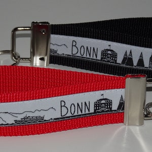 Könnte beinhalten: Zwei Schlüsselanhänger mit schwarzen und roten Stoffbändern. Jedes Band hat einen weißen Streifen mit einer schwarz-weißen Illustration der Bonner Skyline und dem aufgedruckten Wort "BONN".