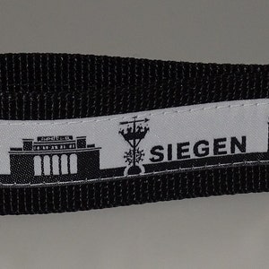 Könnte beinhalten: Schwarz-weißes Schlüsselband mit der Skyline von Siegen, Deutschland. Das Band hat eine schwarze Webstruktur mit einem weißen Band, das die Silhouette der Stadt und den Namen "Siegen" zeigt. Enthält einen silberfarbenen Metallverschluss und einen Schlüsselring.