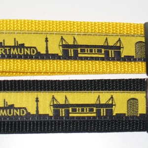 Pode incluir: Dois chaveiros pretos e amarelos com uma silhueta preta do horizonte e do estádio de Dortmund. Os chaveiros têm um anel de metal prateado.