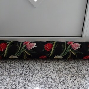 Op de afbeelding: Een lange, cilindrische tochtstrip met een zwarte achtergrond en een bloemenpatroon van rode en roze tulpen met groene bladeren. De stopper is geplaatst aan de basis van een witte deur.