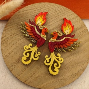 Puede incluir: Dos adornos decorativos de fénix. Cada fénix tiene un cuerpo rojo, alas naranjas y doradas y una cola amarilla. Los fénix se colocan sobre un fondo de madera y un telón de fondo naranja. Los fénix están diseñados con detalles intrincados.