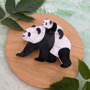 May include: A black and white panda bear brooch with a smaller panda riding on its back. The brooch is set on a round wooden base, with green leaves in the background. The panda brooch is a unique accessory.