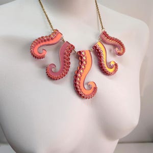 May include: A unique necklace featuring five stylized octopus tentacles in shades of pink, orange, and yellow. The tentacles are attached to a delicate gold-colored chain, creating a striking and whimsical accessory. The necklace is displayed on a white mannequin.