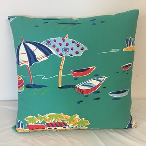 May include: A teal blue throw pillow with a beach scene design. The pillow features two colourful umbrellas, boats, and palm trees.