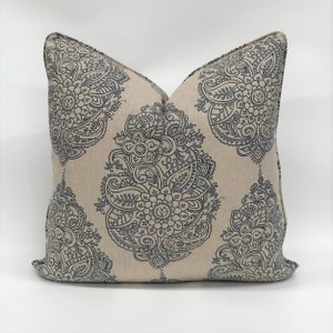 Pode incluir: Uma almofada bege com um padrão paisley azul. A almofada tem formato quadrado e um tecido macio e texturizado.
