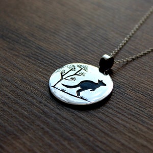May include: A silver necklace with a round pendant featuring a black silhouette of a kangaroo hopping next to a small tree. The pendant is on a silver chain.