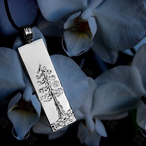 Könnte beinhalten: Eine silberne Halskette mit einem detaillierten Baumdesign. Der Anhänger ist rechteckig und hängt an einem schwarzen Lederband. Der Anhänger wird vor einem Hintergrund aus weißen Orchideenblüten gezeigt.