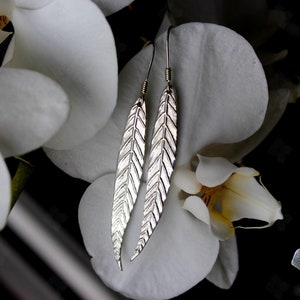 Puede incluir: Pendientes de plata con forma de pluma, con detalles grabados, colgados frente a flores de orquídeas blancas. Los pendientes tienen una forma larga y delgada. El fondo es una mezcla de blanco y negro.