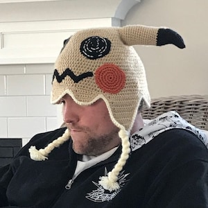 Könnte beinhalten: Eine Person trägt eine gehäkelte braune und beigefarbene Mütze mit schwarzen und orangen Details. Die Mütze ist in Form eines Comic-Charakters mit einem langen, spitzen Ohr geformt. Die Person trägt einen schwarzen Hoodie mit einem weißen Grafikdesign.