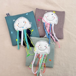 Puede incluir: Tres juguetes de medusas hechos a mano con caras sonrientes y tentáculos colgantes. Cada medusa está unida a una tela cuadrada y texturizada en tonos de verde azulado, gris y verde. Los tentáculos son de varios materiales y colores.