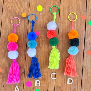 Può includere: Quattro portachiavi colorati con pompon e nappe, etichettati A, B, C e D. Ogni portachiavi presenta una combinazione di colori diversa: rosa, arancione, bianco e blu; blu, bianco e blu; rosso, verde e bianco; e arancione, nero e blu.
