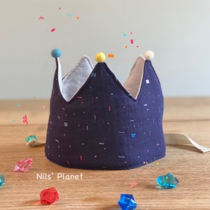 Könnte beinhalten: Eine dunkelblaue Stoffkrone mit weißem Rand und buntem Konfetti-Druck. Die Krone hat einen gelben Bommel oben und weiße Bommel an den Seiten. Die Krone liegt auf einer Holzoberfläche mit bunten Edelsteinen, die um sie herum verstreut sind. Der Text "Nils' Planet" ist unter der Krone sichtbar.