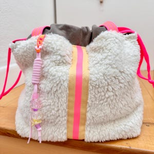 Könnte beinhalten: Ein weißer Sherpa-Beutel mit Kordelzug, der einen vertikalen beigen Streifen mit einem leuchtend neonpinken Mittelstreifen aufweist. Die Tasche hat neonpinke Kordelzüge und einen dekorativen Anhänger mit rosa, lila und transparenten Kunststoffperlen.