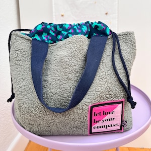 Könnte beinhalten: Ein grauer Kordelzugbeutel aus Sherpa-Fleece mit dunkelblauen Stoffgriffen und einem Kordelzugverschluss. Das Innenfutter zeigt ein Tiermuster in Petrol, Lila und Schwarz. Ein quadratischer Aufnäher in Pink und Schwarz trägt die Aufschrift, let love be your compass.