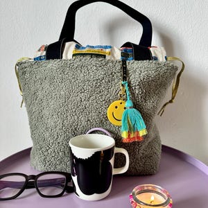 Op de afbeelding: Een grijze sherpa tote bag met zwarte hengsels en een trekkoordsluiting met prints van boekruggen. Een gele smiley-bedel en een veelkleurige kwast hangen aan de tas. Een zwart-witte mok, een zwarte bril en een kaars met kralen zijn aanwezig.