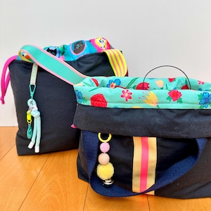 Op de afbeelding: Twee zwarte stoffen tassen met kleurrijke accenten. De voorste tas heeft een turquoise voering met bloemenpatroon, een verticale roze en gouden streep en een kralenbedel. De achterste tas heeft een voering met patroon en een turquoise en gele riem.