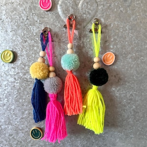 Könnte beinhalten: Eine Sammlung farbenfroher, handgefertigter Quasten mit Holzperlen und Pompons. Die Quasten sind in Blau, Pink, Orange, Gelb und Grau gehalten. Smiley-Magneten sind ebenfalls zu sehen.
