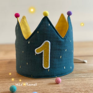 Könnte beinhalten: Eine teal-farbene Stoffkrone mit einer goldglitzernden Zahl 1 Applikation. Die Krone hat eine gelbe Innenseite und ist mit goldenen Flecken und bunten Pompons verziert. Der Text "Nils' Planet" ist unten sichtbar.