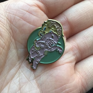 May include: A gold-toned enamel pin featuring a cartoon illustration of a person riding a small, pink, furry creature. The pin is set against a green background.