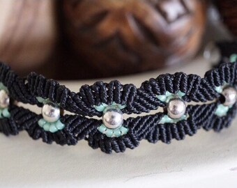 Pode incluir: Um bracelete de macramé preto com contas prateadas e detalhes em azul claro.