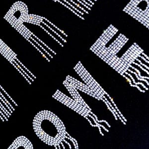 Può includere: Una maglietta nera con le parole "Girl Power" scritte in strass argentati con fili di strass pendenti.