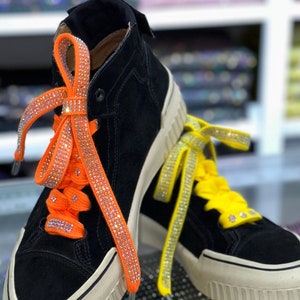 Puede incluir: Un par de zapatillas altas negras con suelas blanquecinas. Las zapatillas tienen cordones naranjas y amarillos con pedrería. Los zapatos tienen una textura similar a la gamuza.