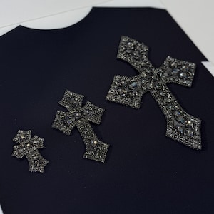 Peut inclure: Trois patchs en forme de croix noire avec des strass sur un fond de tissu noir.