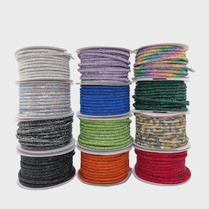 May include: A collection of twelve spools of round, textured cord arranged in three vertical columns. The cords feature various colors including silver, purple, rainbow, blue, green, dark gray, orange, red, and multi-colored metallic finishes.