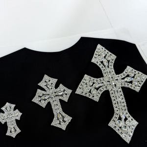 May include: Black t-shirt with three rhinestone cross designs. The crosses are different sizes and are all made of clear rhinestones.
