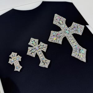 Pode incluir: Três patches de ferro em forma de cruz prateados com strass iridescentes. As cruzes são de tamanhos diferentes, com a cruz maior na parte superior e a cruz menor na parte inferior.