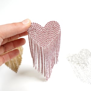 May include: A pink rhinestone heart-shaped earring with dangling fringe. The earring is held up against a white background. Another gold-colored heart-shaped earring is visible in the background.