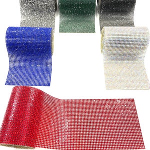K&ouml;nnte beinhalten: Rollen mit glitzerndem Strassband in verschiedenen Farben, darunter Silber, Gr&uuml;n, Schwarz, Blau, Klar und Rot. Das Band ist auf einer wei&szlig;en Oberfl&auml;che ausgerollt.