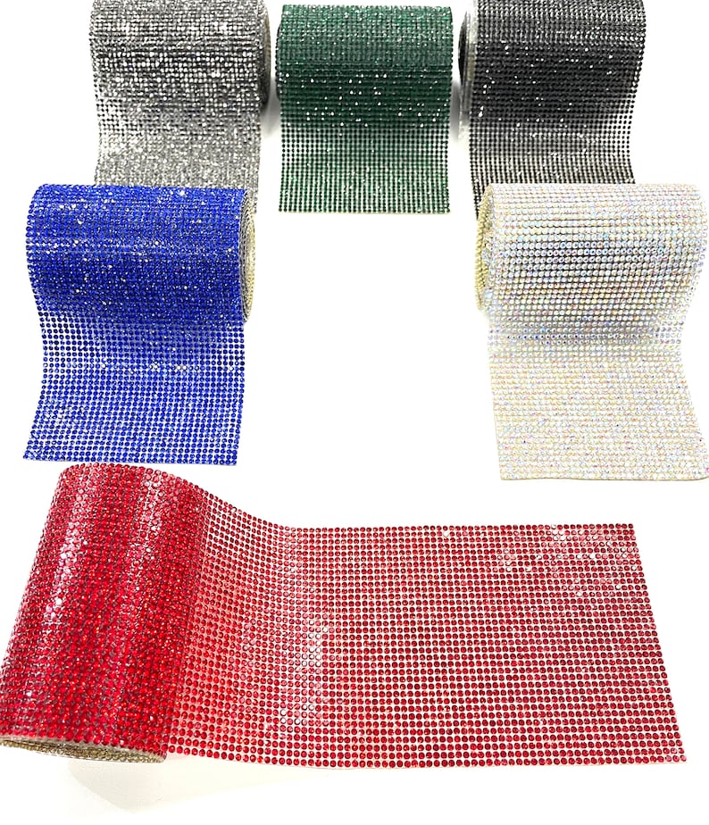 K&ouml;nnte beinhalten: Rollen mit glitzerndem Strassband in verschiedenen Farben, darunter Silber, Gr&uuml;n, Schwarz, Blau, Klar und Rot. Das Band ist auf einer wei&szlig;en Oberfl&auml;che ausgerollt.