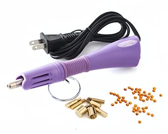 May include: Purple electric hot fix tool with a black cord and plug. The tool is used to attach rhinestones to fabric. The image also shows a clear plastic circle, a handful of gold-colored metal tips, and a scattering of orange rhinestones.