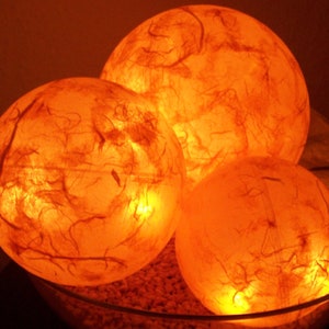 Könnte beinhalten: Drei runde, orangefarbene Papierlaternen mit einer strukturierten, faserigen Oberfläche. Die Laternen werden von innen beleuchtet und erzeugen ein warmes Leuchten.