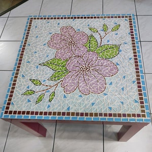 Puede incluir: Una mesa cuadrada de mosaico con un diseño floral. La mesa está hecha de pequeñas baldosas de colores y presenta dos flores rosas con hojas verdes.