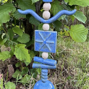 May include: A vertical garden stake featuring stacked ceramic elements in blue and off-white. The design includes a pointed top, a curved horizontal piece, a square tile with a star-like relief, cylindrical segments, and a large blue sphere base.