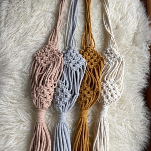 Könnte beinhalten: Vier Makramee-Pflanzenaufhänger in Rosa, Grau, Senfgelb und Creme. Jeder Aufhänger hat ein gewebtes Korbmuster mit einer langen Quaste. Die Aufhänger werden auf einer flauschigen, weißen Oberfläche präsentiert.