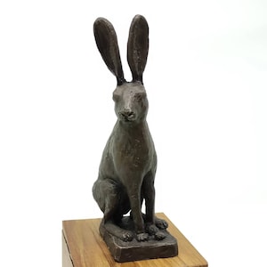 Pode incluir: Uma escultura de bronze de uma lebre sentada com orelhas compridas. A lebre está olhando para a frente com a cabeça ligeiramente inclinada para baixo. A escultura está sobre uma base de madeira.