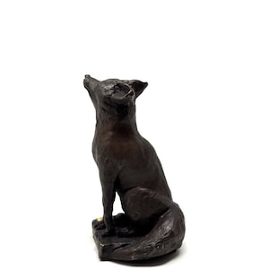 May include: A bronze fox sculpture in a seated pose, gazing upwards. The fox is detailed with visible fur texture and sits on a small, square base. The sculpture has a dark, antique finish.