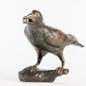 Puede incluir: Una escultura de bronce de un cuervo posado en una roca, sosteniendo un pequeño objeto en su pico.