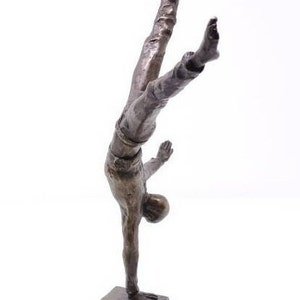Può includere: Scultura in bronzo di una persona in equilibrio sulle mani sopra una pila di libri. La scultura è su una base circolare.