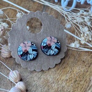 Könnte beinhalten: Zwei runde Ohrstecker mit floralem Muster. Die Ohrringe haben einen schwarzen Hintergrund mit rosa, blauen und grünen Blumenmustern. Sie werden auf einer braunen Karte mit gewelltem Rand präsentiert, mit getrockneten Blumen im Hintergrund.